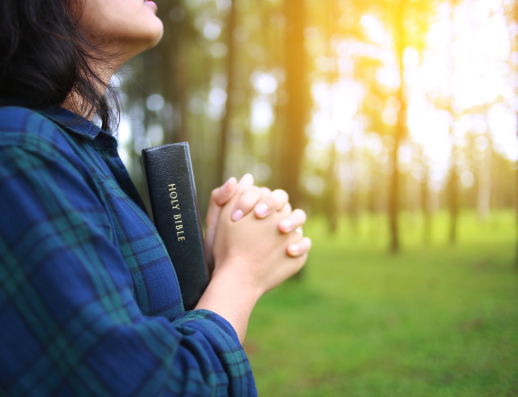 sun shiny day in background outside, woman holding Bible and praying  5 Things Prayer Does in the Life of a Christian: Matthew 6:9-13